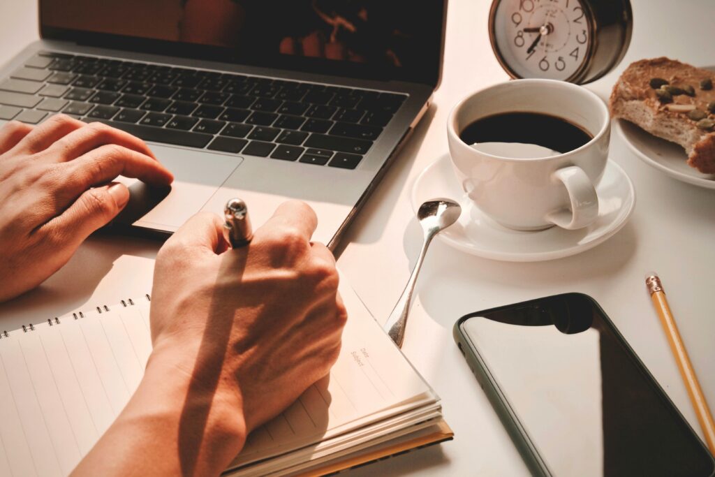 Close-up of hands writing in a notebook with coffee, laptop, smartphone, and alarm clock nearby.